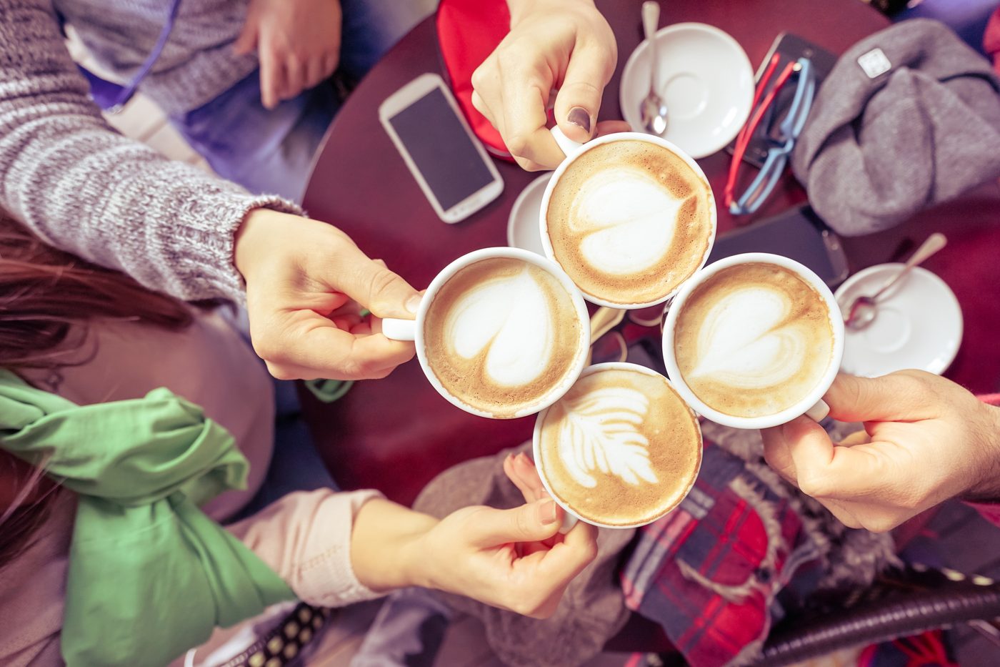 Four friends holding coffee cups together over a cafe table, shot from above showing all hands
