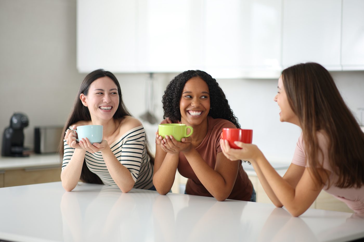 Three roommates laughing over colourful mugs in a bright kitchen